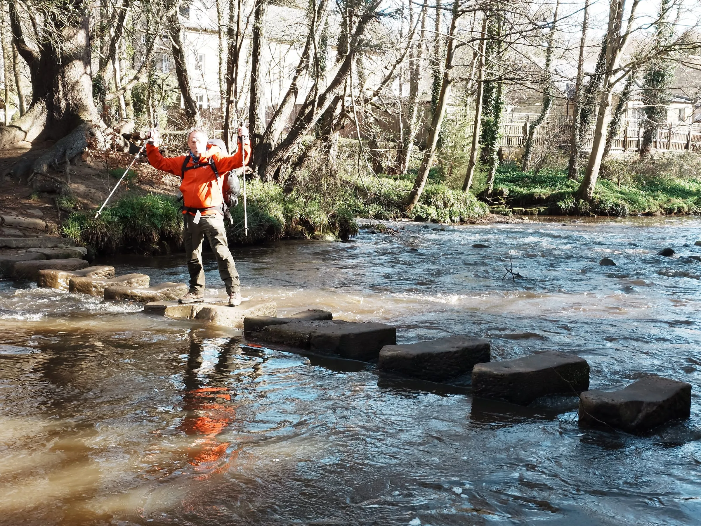 Toby crossing a river on stepping stones, pack on, poles out